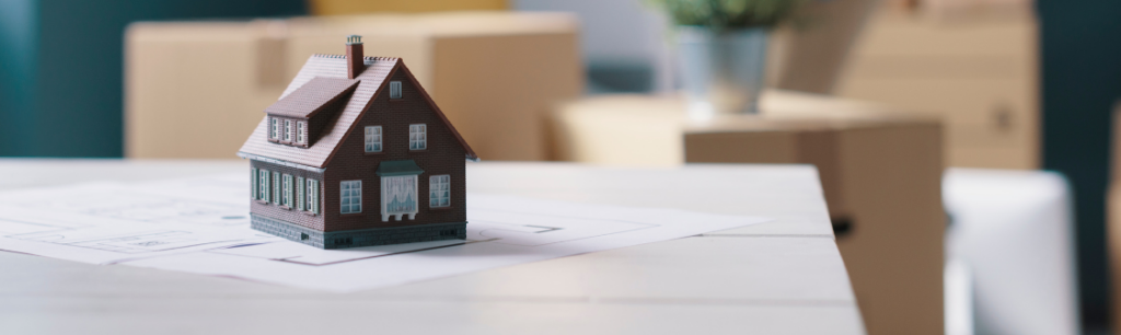 A toy house sits on a table, while man moves boxes in the background.